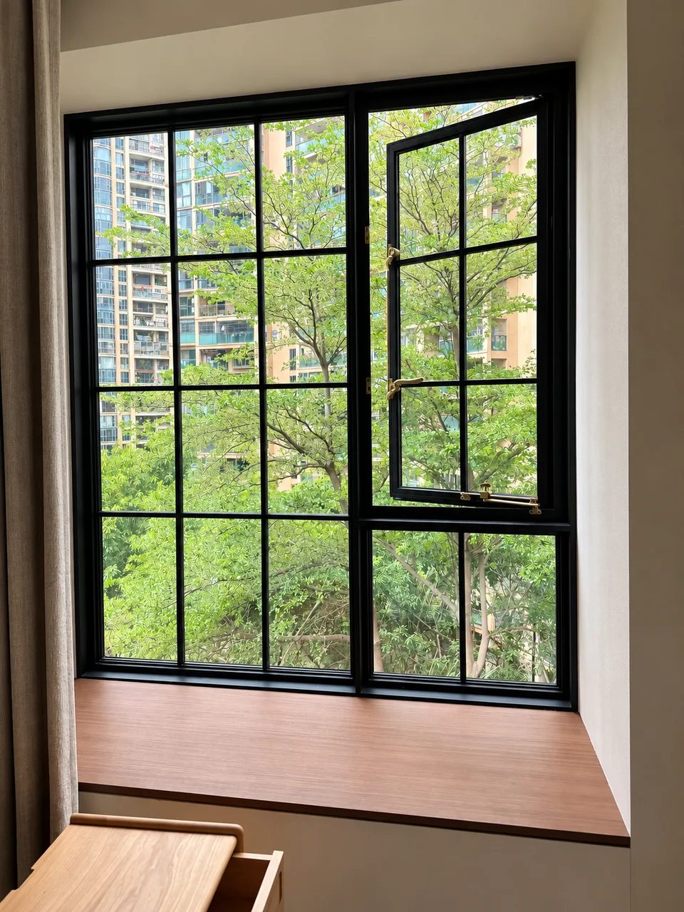 Black French grid casement window with brass-tone handles overlooking leafy trees